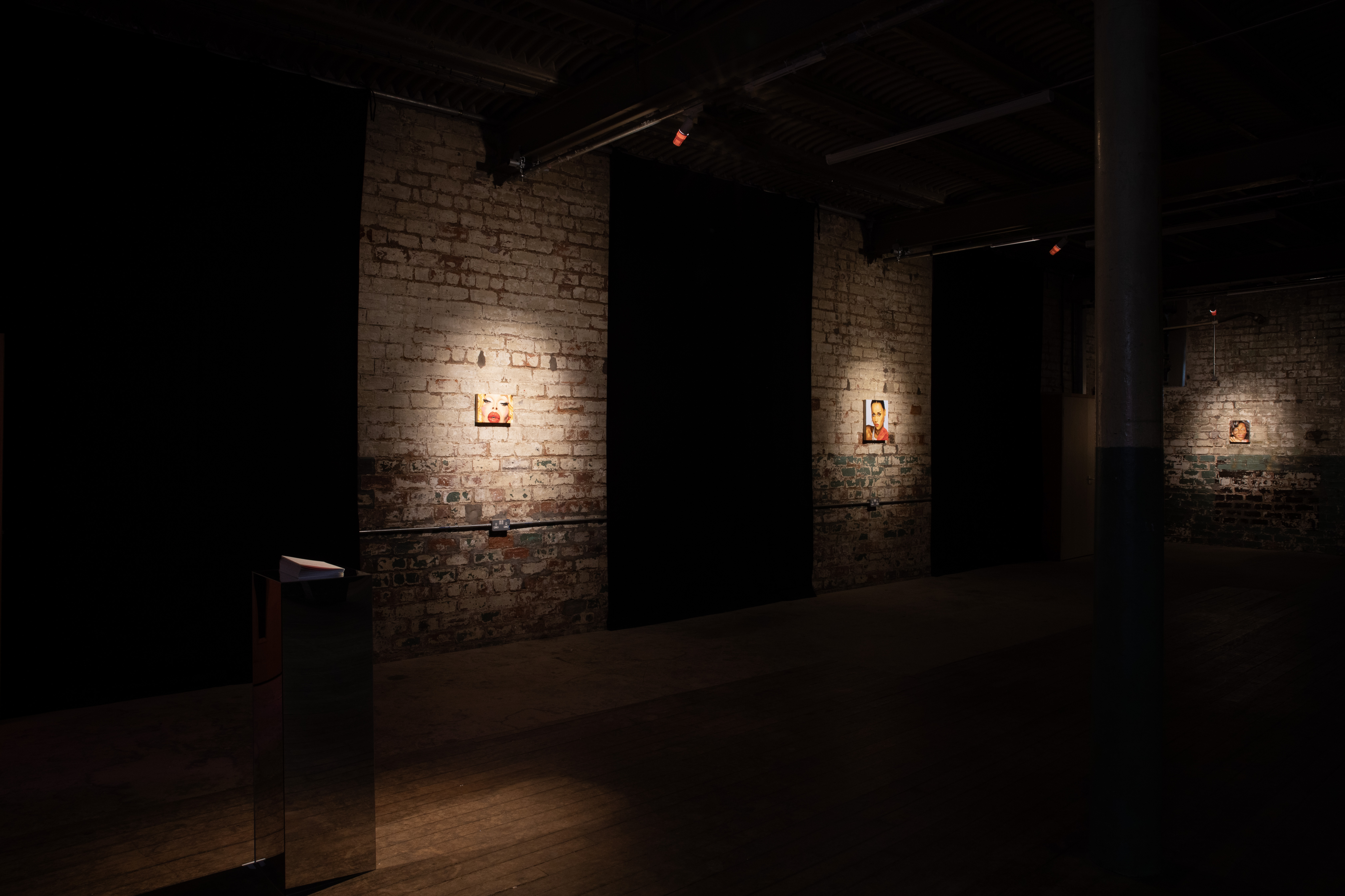 Dark gallery space with exposed brick pillars and walls. Two small spotlit portrait paintings of faces are visible on separate wall sections, with a mirrored plinth holding papers in the foreground and a third painting faintly lit in the distance.