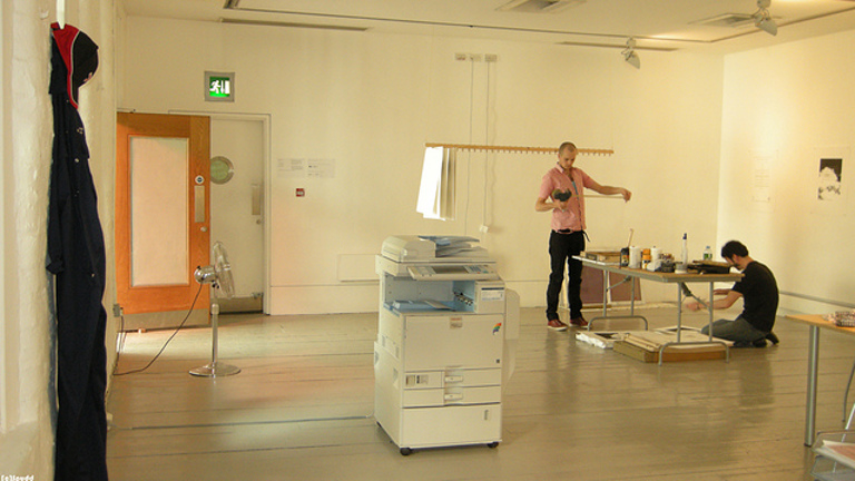 two people in a room fiddling with a table, in the centre of the room is an office printer.