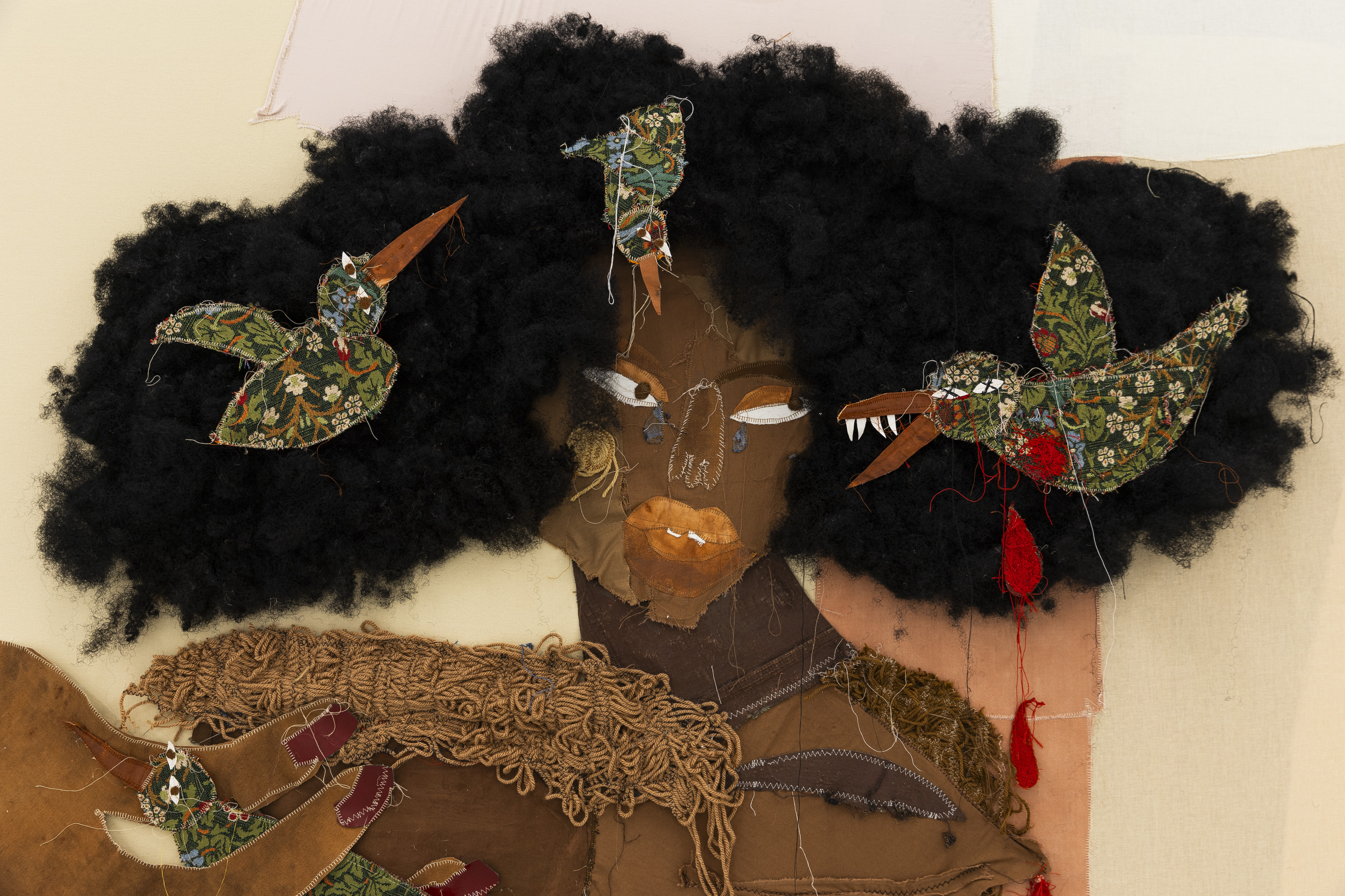 Close-up detail of a mixed-media textile portrait showing a face with a fierce expression, teary glass-bead eyes, and patterned fabric hummingbirds with sharp fangs and trailing red thread nesting in voluminous black woollen hair.