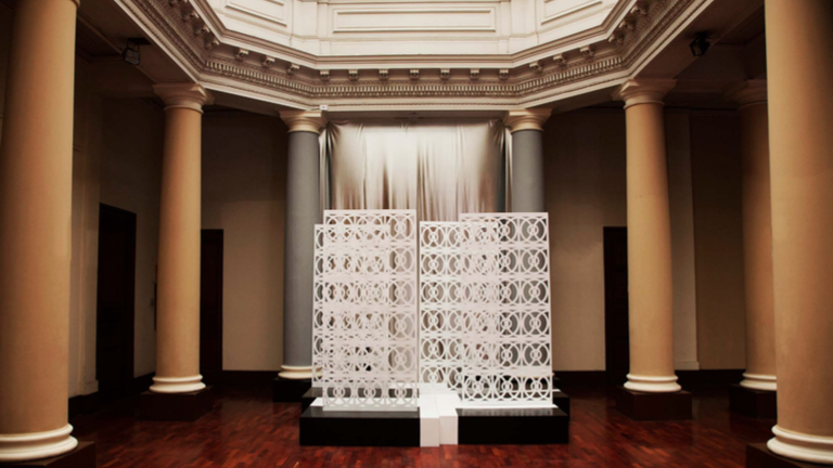 An interior of a hexagonal room with columns. A sculptural object is exhibited in the  middle 