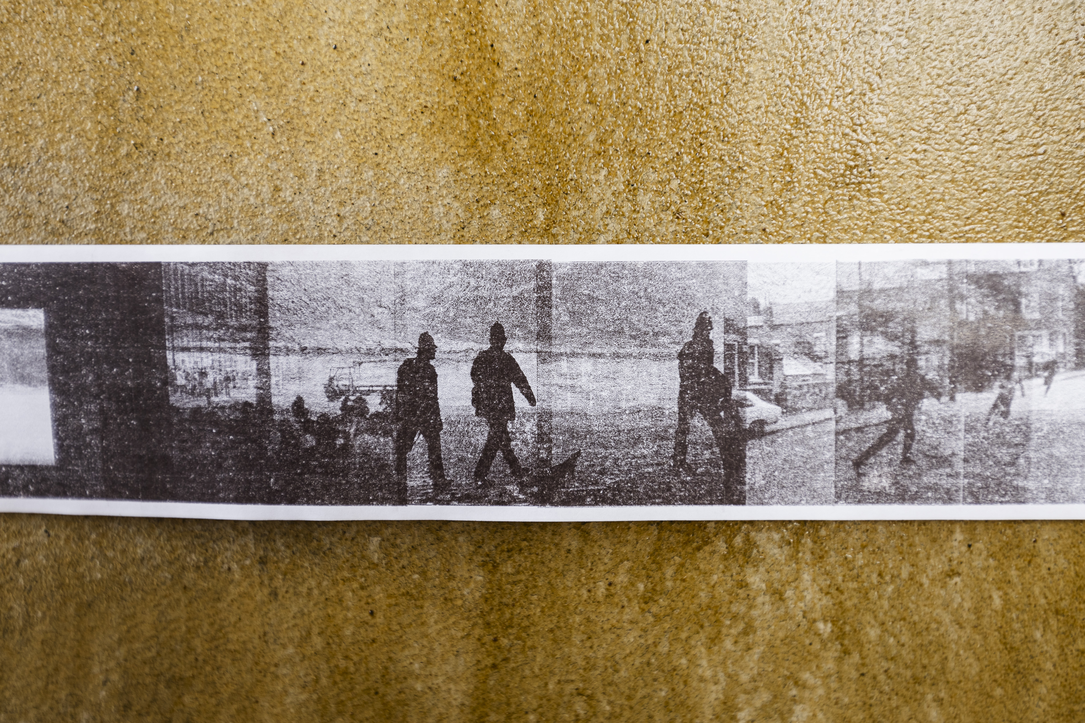 Close-up of a printed photographic strip taped to a mustard-yellow textured wall. The grainy black-and-white images show silhouetted figures walking along a street or waterfront, captured in sequence like film stills or a contact sheet.
