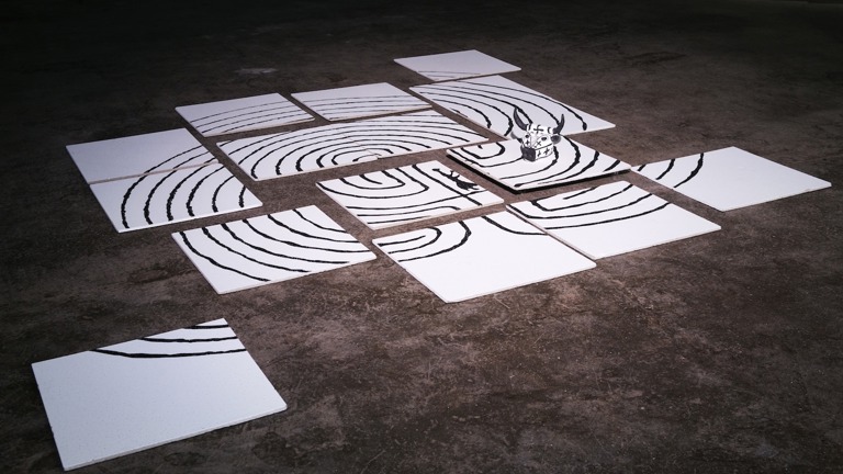 A series of white panels laid out on a floor. The panels have a series of black lines painted on them in concentric circles. A small paper model of a bull's head is also visible  