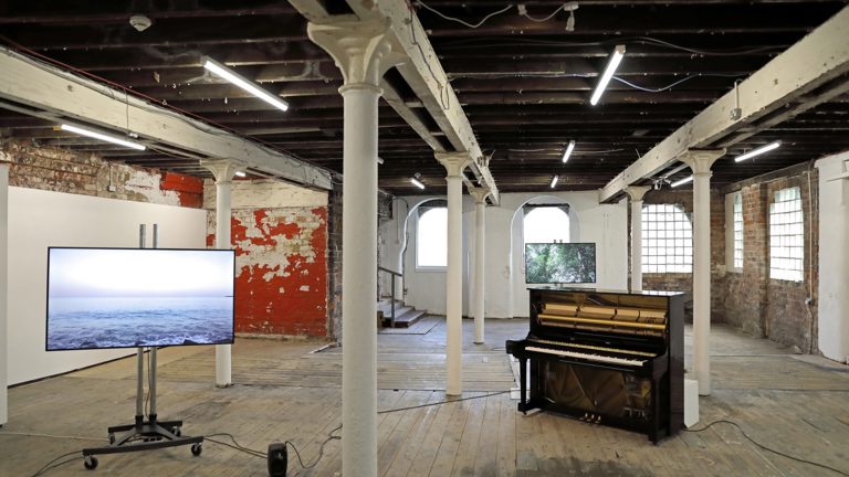 An installation of Yuko Mohri's work in the Pipe Factory, Glasgow. There is a piano and tv screen on a stand. The screen shows an image of the ocean, the piano is wired up to speakers.