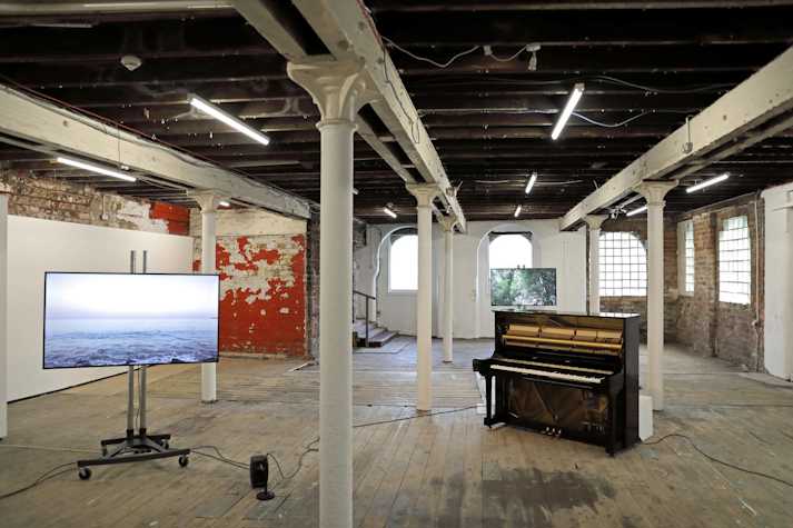 An installation of Yuko Mohri's work in the Pipe Factory, Glasgow. There is a piano and tv screen on a stand. The screen shows an image of the ocean, the piano is wired up to speakers.