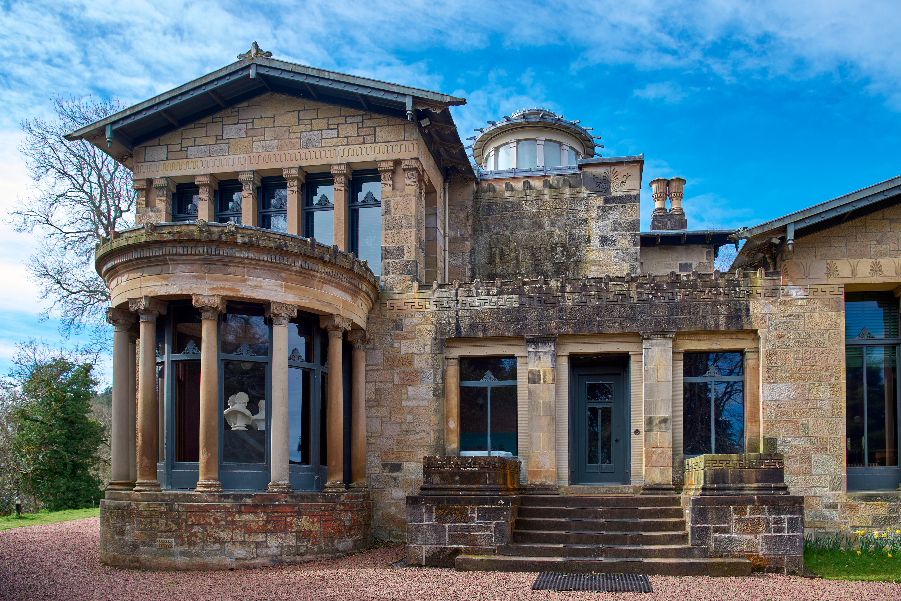 A three-quarter view of Holmwood House's sandstone facade, showing its columned rotunda, domed lantern, and chimneys against a bright sky.