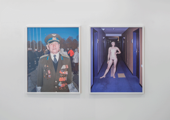 Two framed photographs are placed sided by side. The one on the left shows an old man dressed in military attire. The one on the right shows a nude woman standing in the hallway of what seems to be a hotel.
