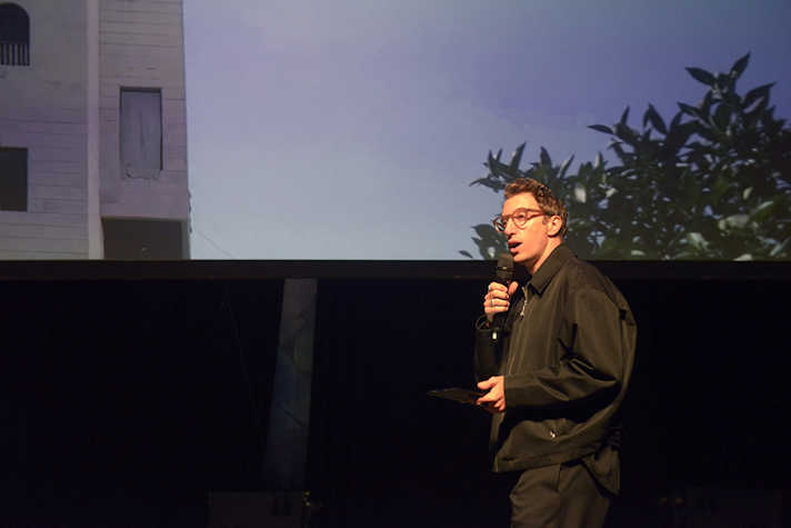 figure with a microphone in front of a screen showing a building and a tree