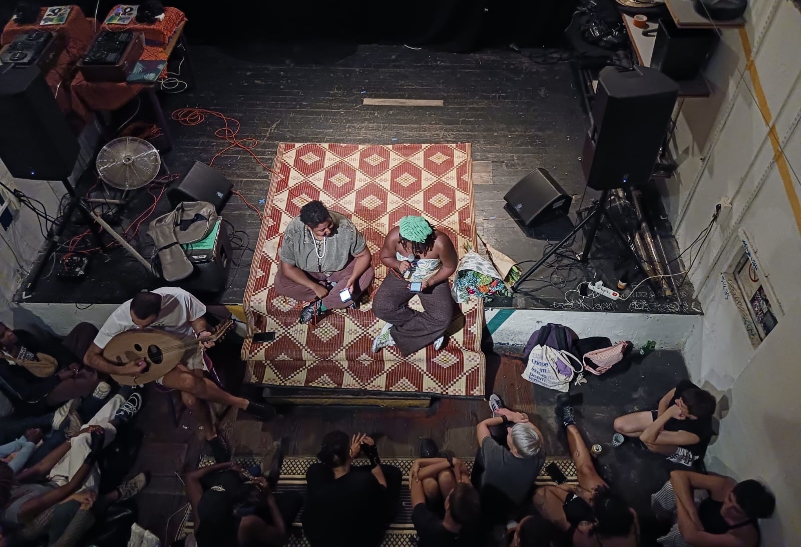 A crowd gathers around two well-lit individuals seated on a red and white patterned rug on a wooden stage. Electronic devices and sound equipment are scatted around them. Both individuals hold their phones, and one reads into a microphone. 