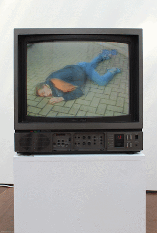 cube monitor on top of a plinth, the screen shows a man lying down on paved floor.
