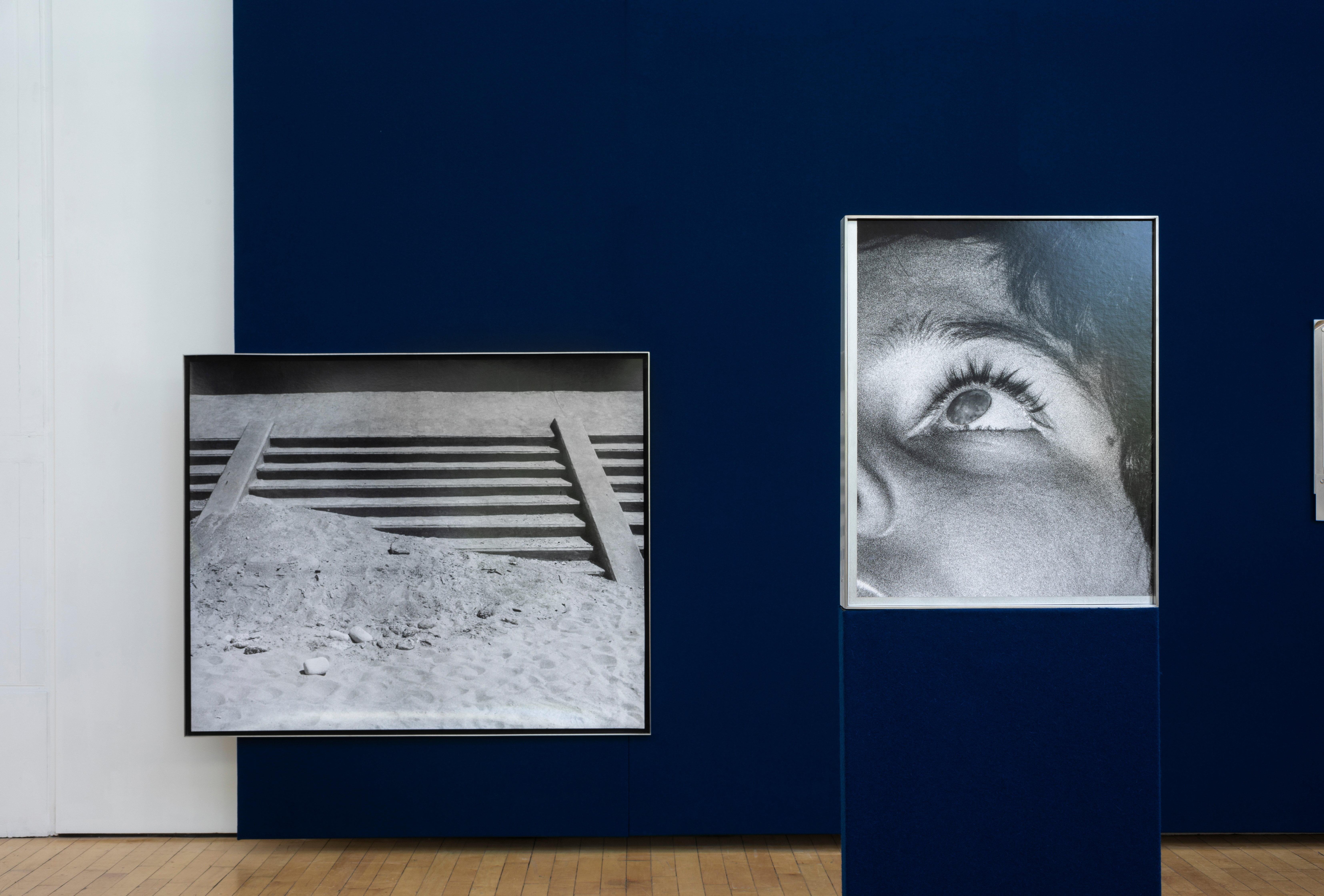 Two black and white photographs mounted on a navy blue gallery wall, one showing wooden slats laid across sand, the other a close-up of an eye with heavy lashes looking upward.