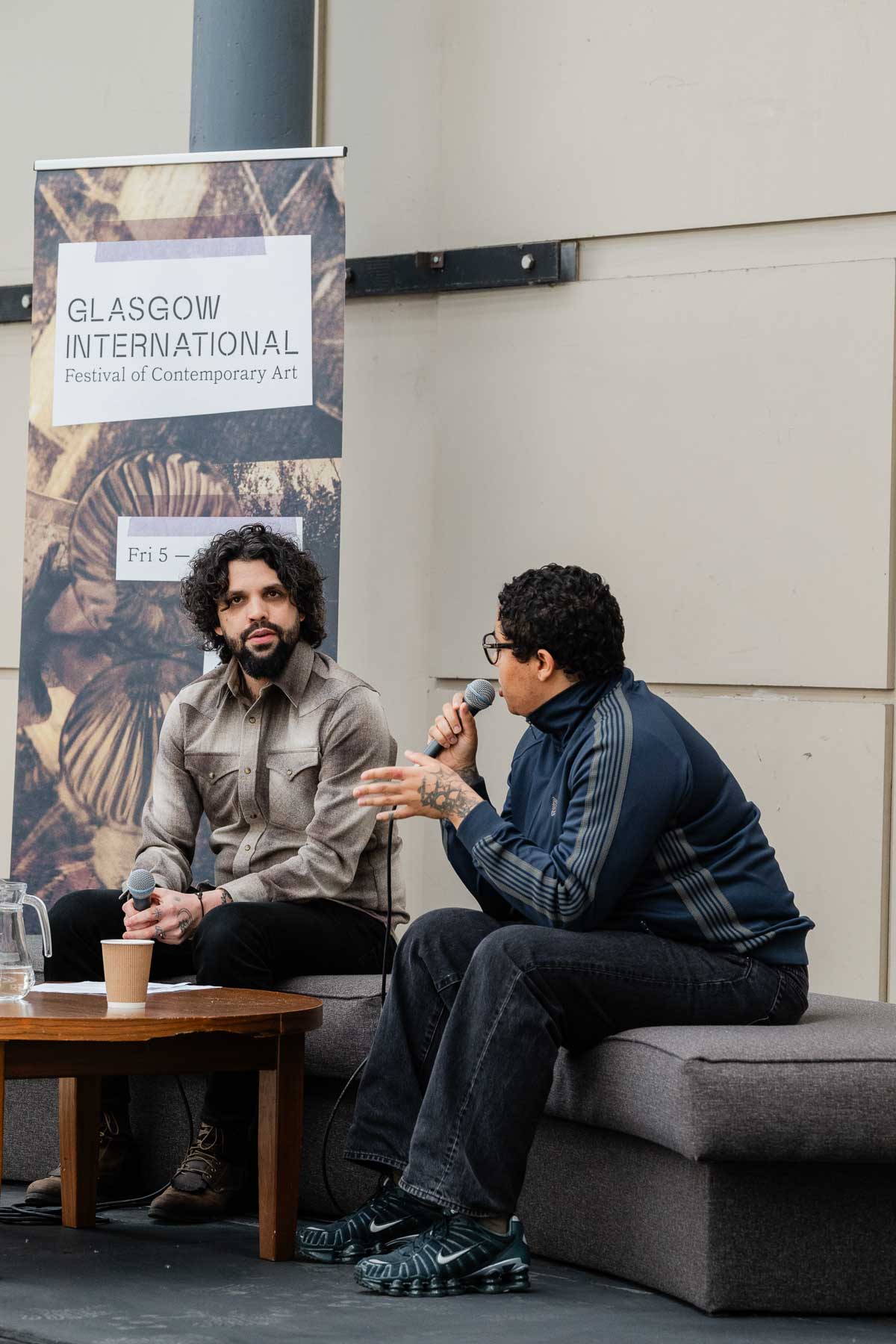 Two people sit talking on a bench-style seat, one gesturing with a microphone in hand and speaking, the other listening. A Glasgow International festival banner stands behind them, and a small table with a coffee cup sits in front.