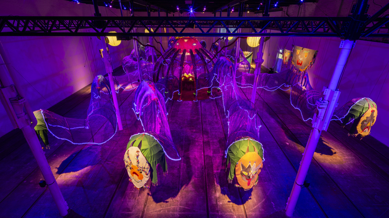 An overhead installation show showing several fabric pieces of green, purple, pinks, and yellows. These sculptural works make the body of an octopus like shape, the end of each tentacle is a green hollowed oval with mask like features. 