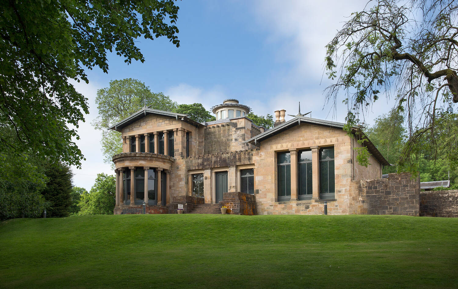The sandstone Greek Revival exterior of Holmwood House, with its columned bay window and rooftop lantern, set on a lawn framed by trees under a blue sky.