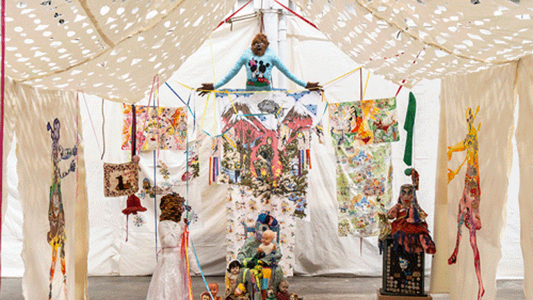 Artwork and sculptures are displayed under a fabric canopy. There is a seated sculpture and a rug in front.