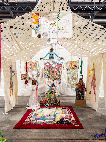 Artwork and sculptures are displayed under a fabric canopy. There is a seated sculpture and a rug in front.