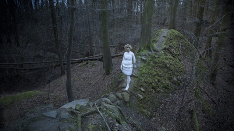 A photograph of a dark woodland. A figure dressed in white clothes is walking along a rocky path