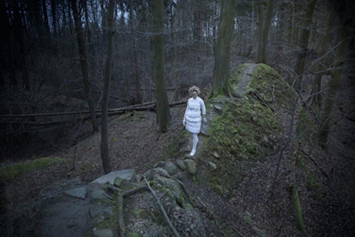 A photograph of a dark woodland. A figure dressed in white clothes is walking along a rocky path