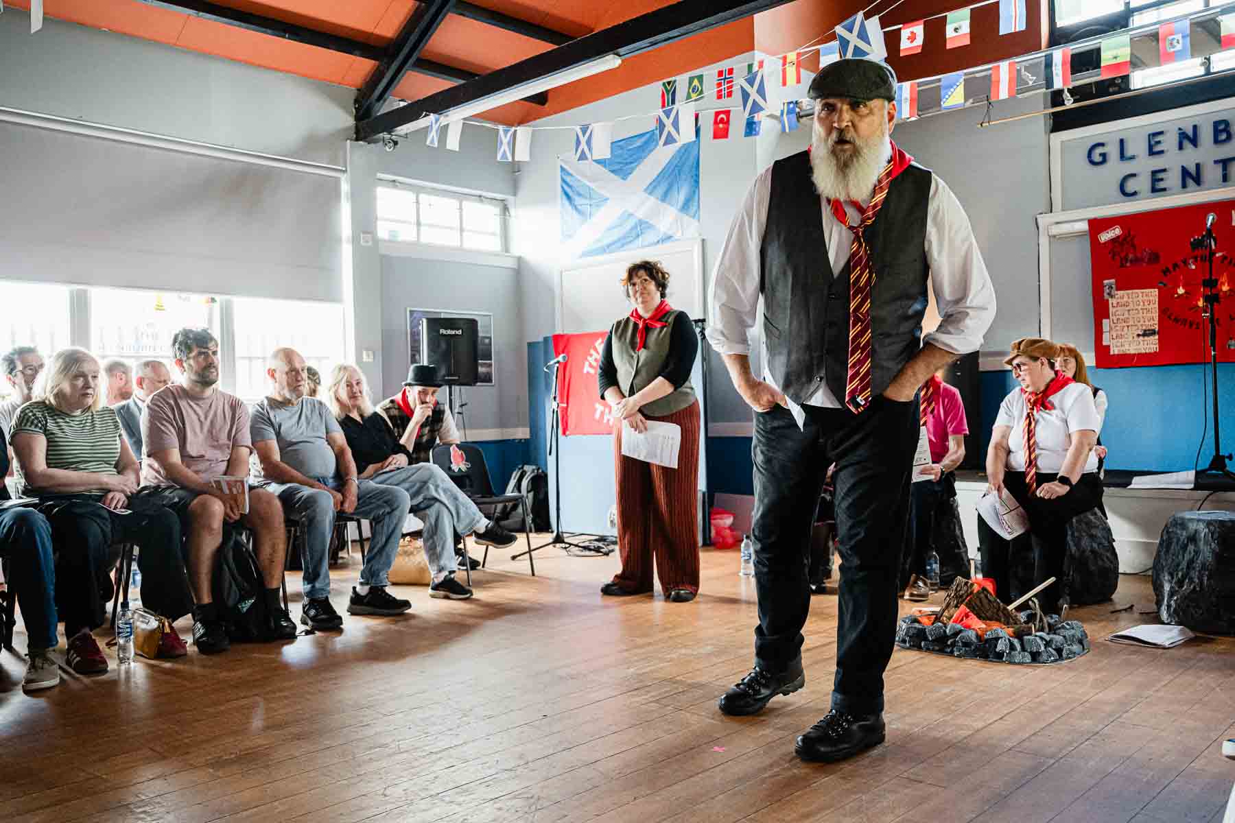A bearded performer in a waistcoat and striped tie strides across the floor mid-performance, watched by seated audience members and a costumed cast member behind him, with a Saltire flag on the wall.