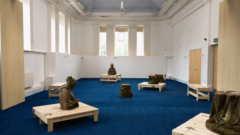 In a large room with period feature ceiling and blue carpet, a series of wooden structures and sections of red sandstone sculptures are spaced out