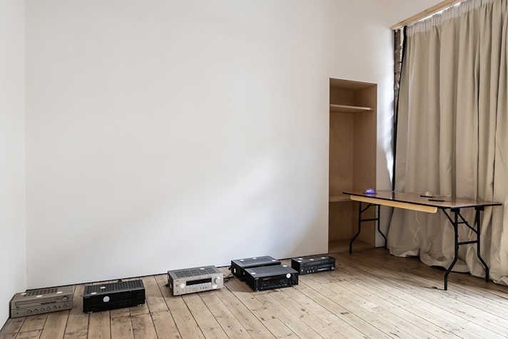 A row of old VHS players and DVD players are displayed on the floor of the room. In the corner of the room there is a table with a phone on it.