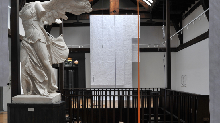 white sheets of fabric hanging from a wood beamed ceiling. their is a winged headless marble statue in front