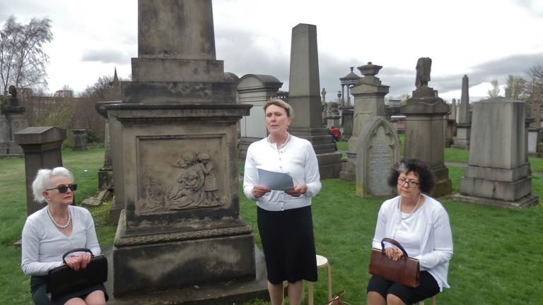 Performance in Glasgow Necropolis. Three women in white cardigans and black skirts and dark purses are sitting down and reading out loud from a paper with tomstones behind them.