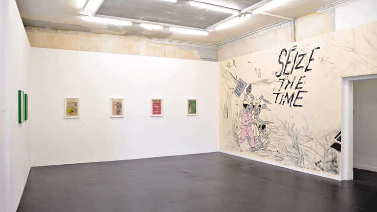 gallery with lithograph prints installed along the left and back walls. the right wall shows a mural with a group of protesters holding a picket sign and guns. mural text reads, 'seize the time'