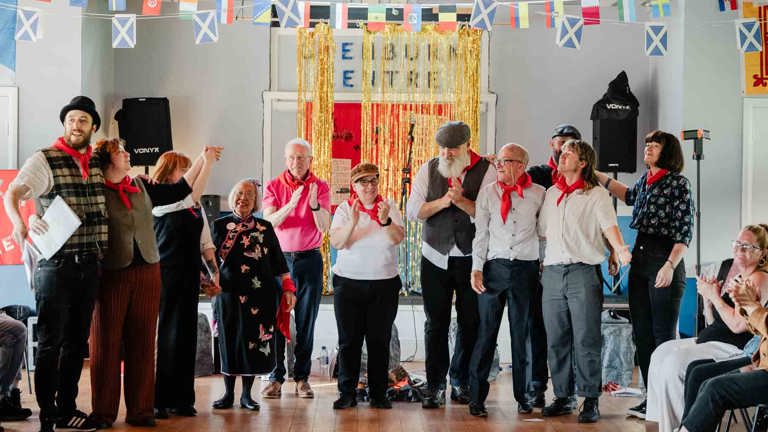 A group of community performers in red neckerchiefs stand arm in arm, applauding, in front of a gold tinsel curtain and international flag bunting at the Glenburn Centre, part of Keira McLean's performance.