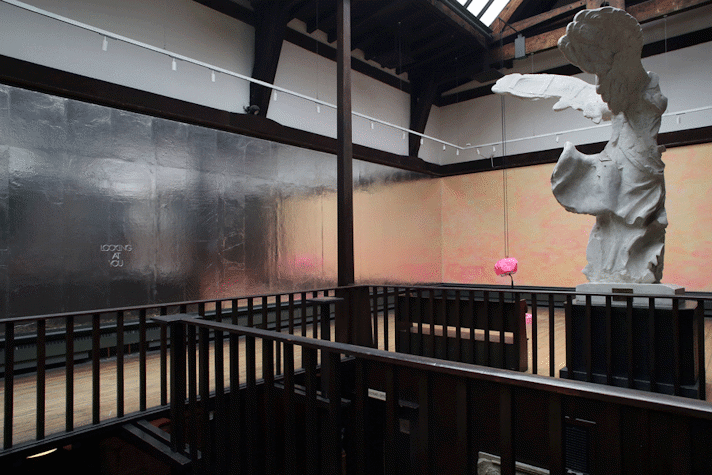 The words "Looking at you" are written on the walls of the hallways, which is covered in metallic paper or paint. There is a angel like sculpture on one side of the hallway and next to it is another pink sculpture hanging off the ceiling.