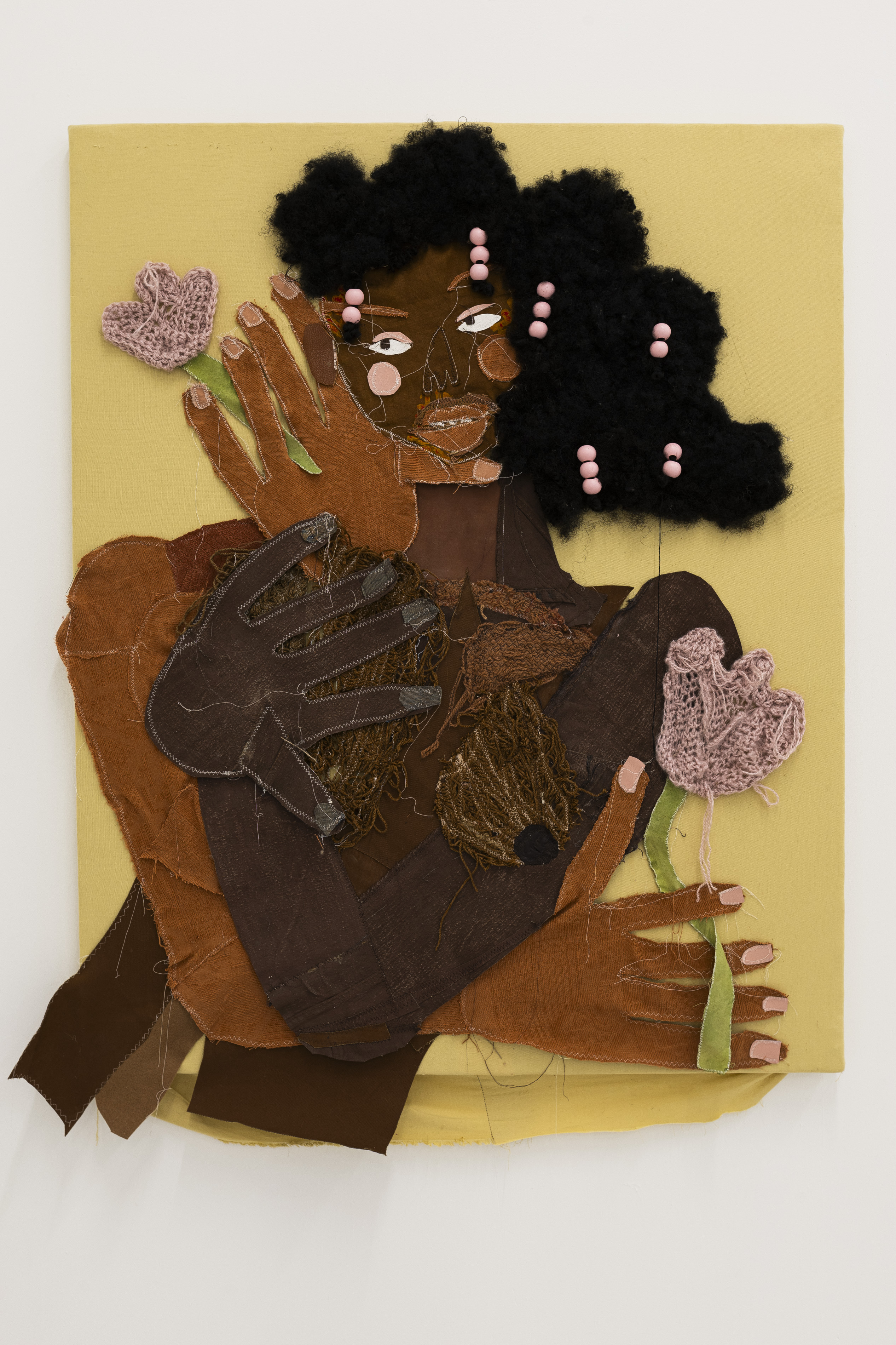 A mixed-media textile portrait of a figure with sculptural black woollen afro puffs studded with pink beads, oversized stitched hands raised beside the face, and knitted pink flowers, set against a mustard-yellow ground.