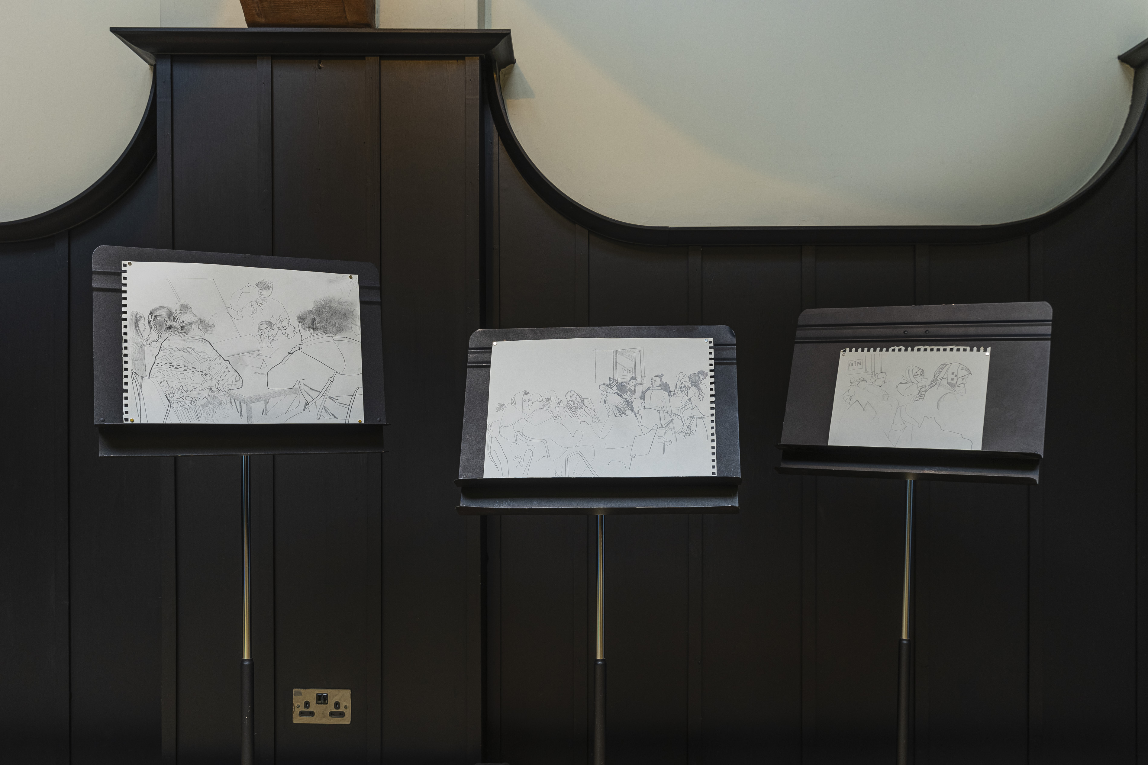 Three music stands hold pencil sketchbook drawings of group scenes, figures at a table and gathered together, displayed against dark wood panelling.