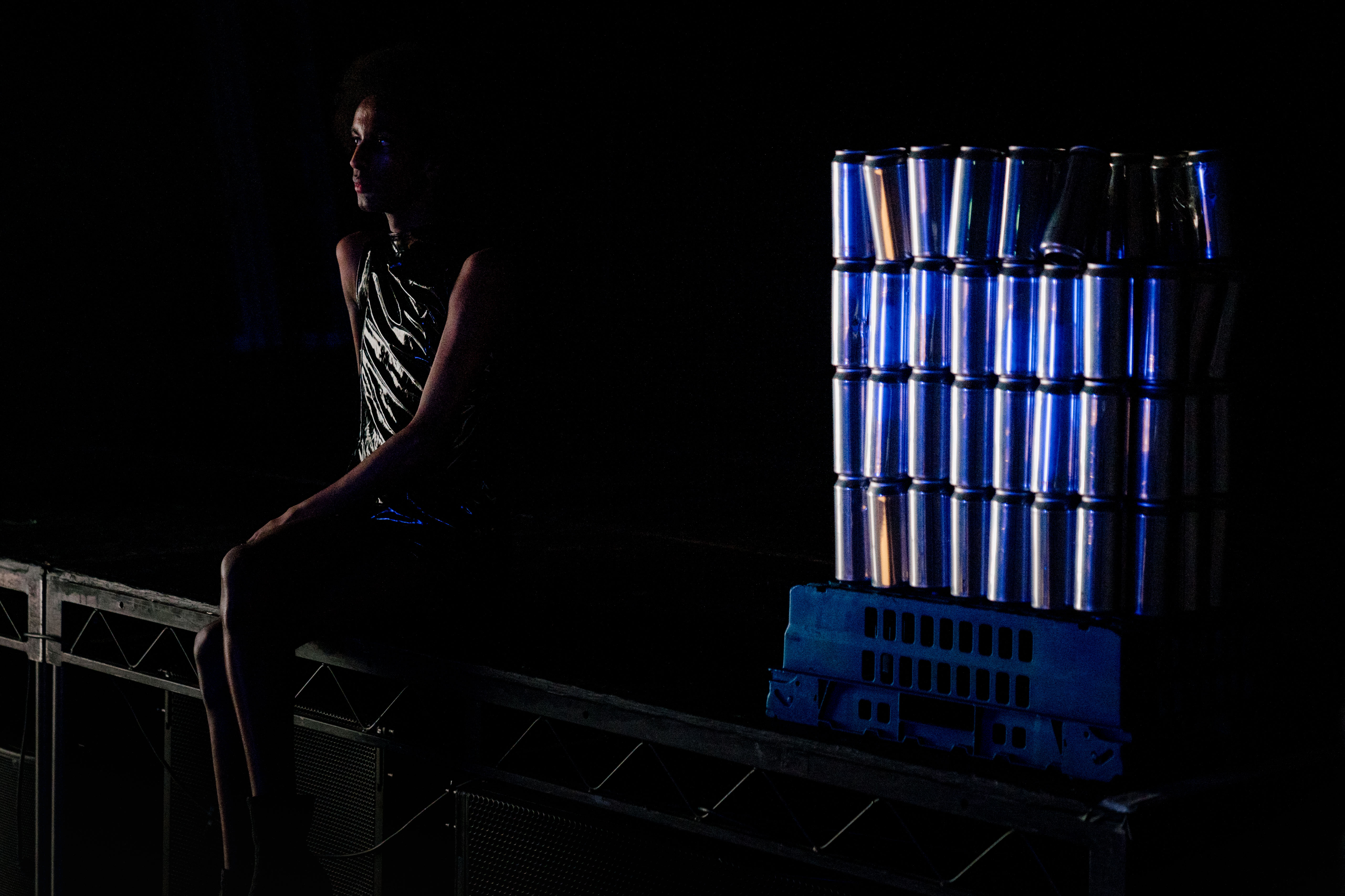 A performer wearing a shiny metallic vest sits on stage in a dark perfromance space. Beside them is an upturned storage tray stacked with layers of cans.