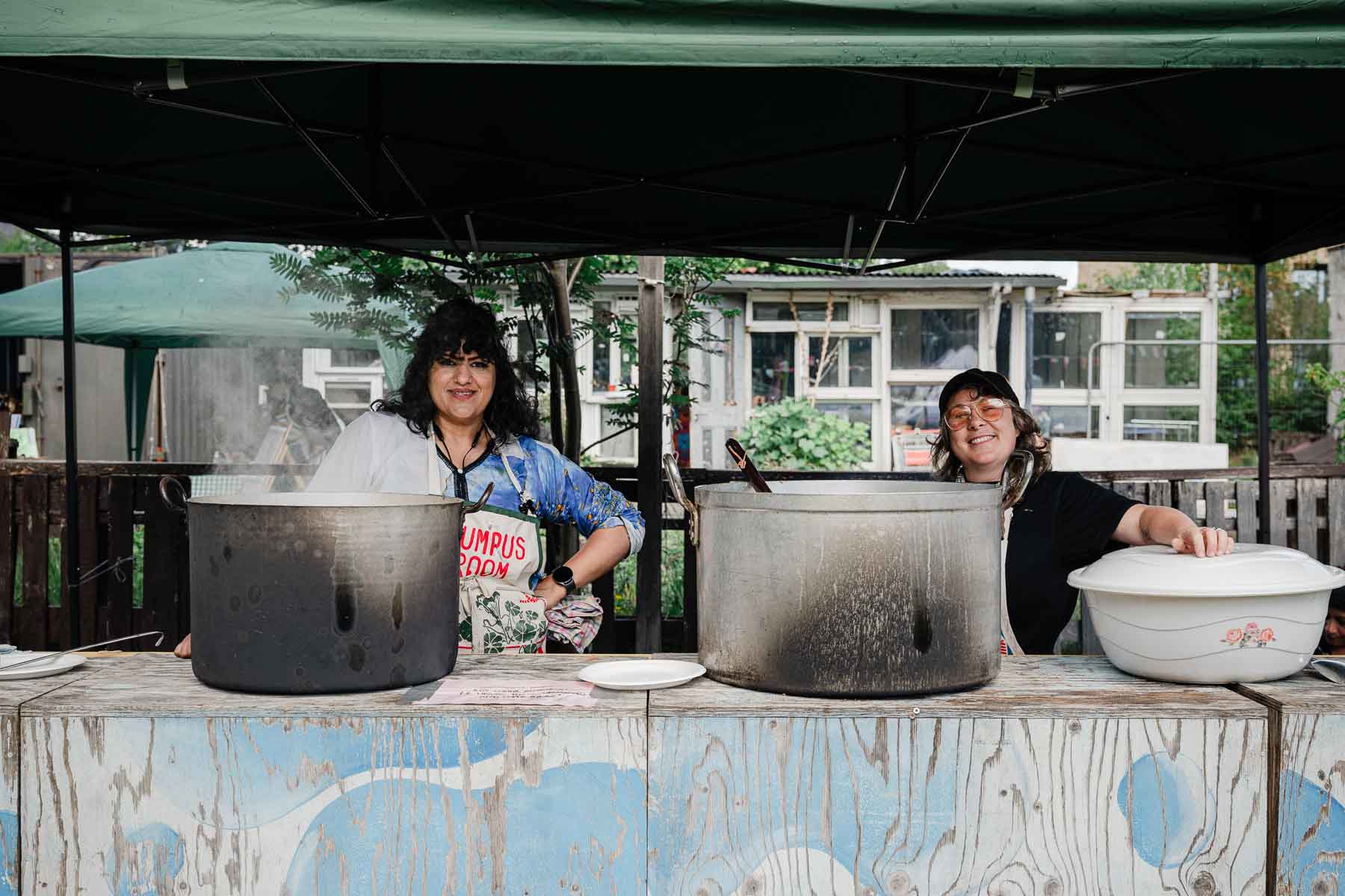Two smiling volunteers stand behind large steaming cooking pots at an outdoor stall with a green gazebo, serving food from a wooden counter painted with a cloud design.