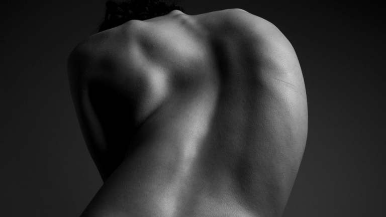 Black-and-white photograph of a person's curved back. The lighting and shadows emphasis the sculptural form of the body.