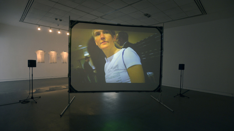 Large screen installed in a white walled gallery with speakers on either side. A film is projected onto the screen. An image of a person is visible on the screen