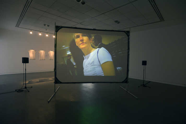 Large screen installed in a white walled gallery with speakers on either side. A film is projected onto the screen. An image of a person is visible on the screen