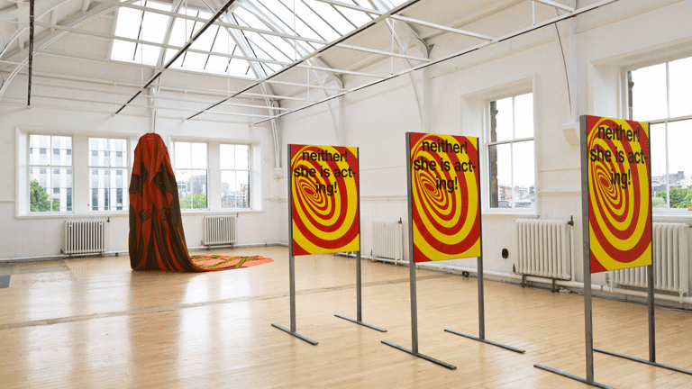 Three identical metal stands hold three identical portrait oriented graphic posters with a red and yellow swirl and black text overlaid. The text reads 'neither! she is acting!'. In the backround a large cape is suspended from the roof and trails along the floor.