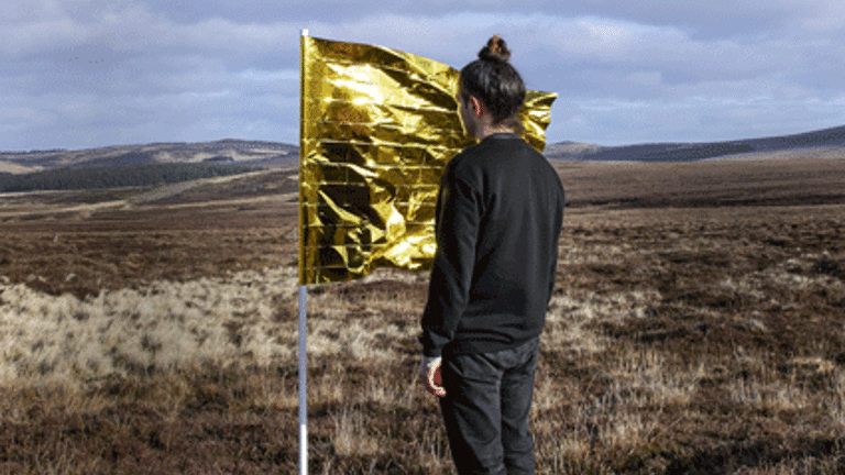 The image shows a figure in black clothes, with brunette hair and topknot, standing with their face close to the fabric of a flag. The flag is made from a gold emergency blanket. The flag and figure are in an open field with hills in the far distance and blue sky.