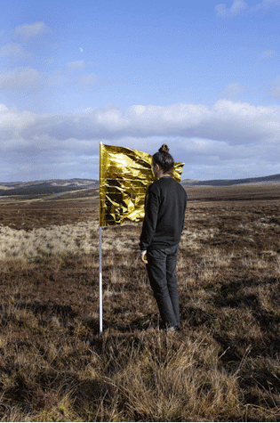 The image shows a figure in black clothes, with brunette hair and topknot, standing with their face close to the fabric of a flag. The flag is made from a gold emergency blanket. The flag and figure are in an open field with hills in the far distance and blue sky.