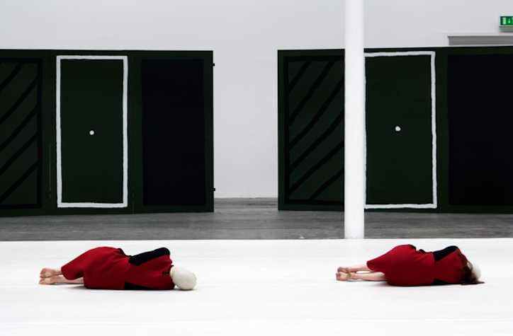 two people dressed in red lying on the floor with their backs faced to us. They're on top of a white floor covering. behind them is two black freestanding displays, each with a white rectangle with a white dot in the centre.
