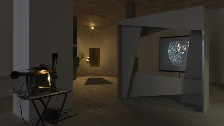  Darkened art gallery with a projected image of a man’s profile over a textured background, an old film projector on a stand, and various abstract installations in the background under soft lighting.