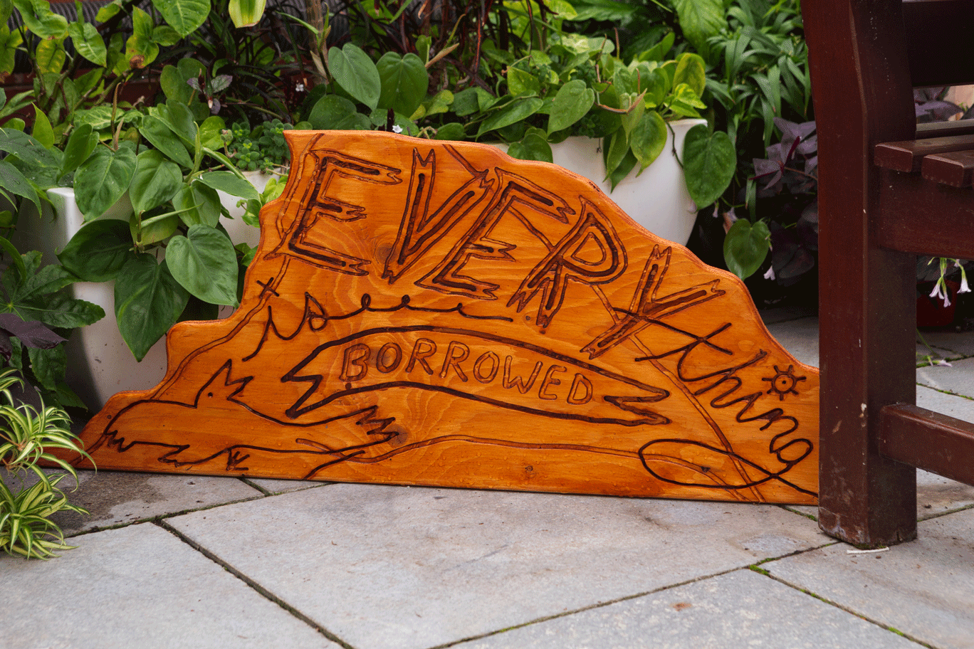 Orange-coloured wooden sculpture with the words Everything Borrowed resting in an environment with plants