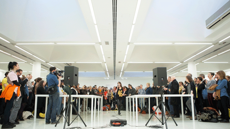 A woman is speaking into a microphone, standing in the centre of a large hall. There is a big crowd standing at a distance, all around her. In front of her, there are two long tables running parallel to each other. On the left there is a man taking a video of the event. Placed ahead of the tables and the man, are two speakers, held up by tripod stands. They are connected to a control box placed between them.