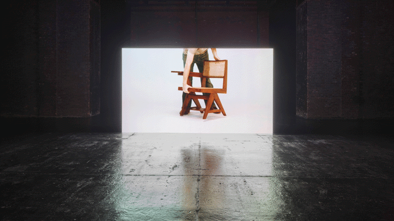 Installation view of a video showing a figure adjusting a chair