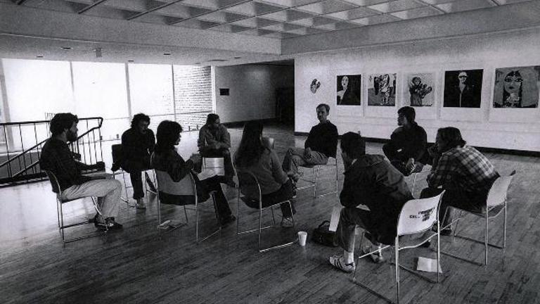 black and white film still showing a group of 9 people sat in chairs having a discussion.