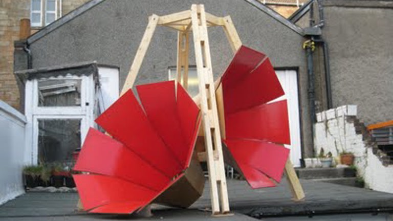 Outdoor art installation featuring a wooden frame supporting two large, curved red panels, resembling an abstract sculpture, set against a background of houses