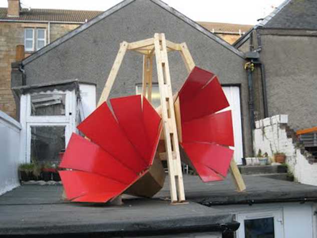 Outdoor art installation featuring a wooden frame supporting two large, curved red panels, resembling an abstract sculpture, set against a background of houses