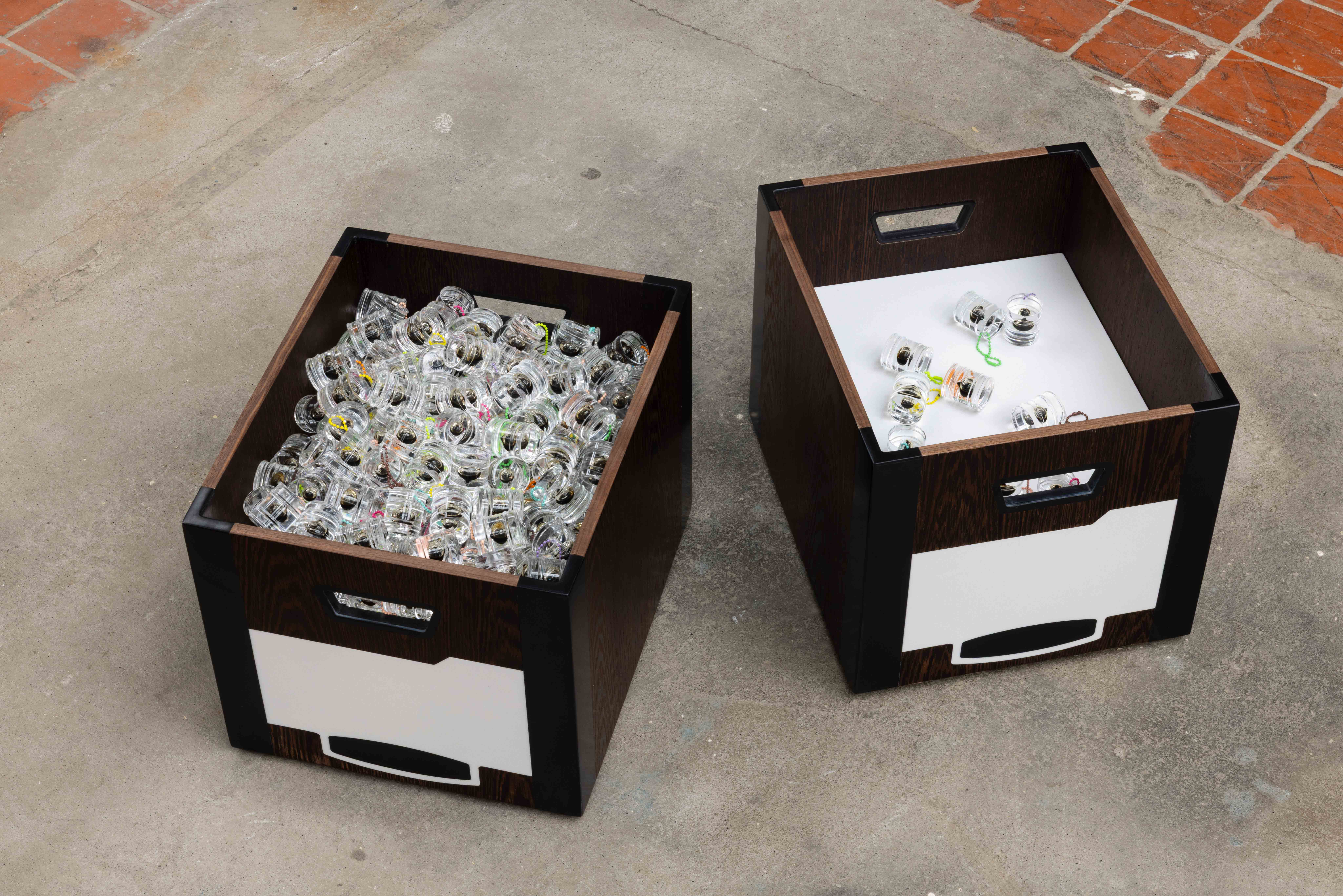Two open wooden storage crates on a concrete floor, one filled with dozens of small glass jars containing beaded charms, the other holding a scattered handful of the same jars on a white base.