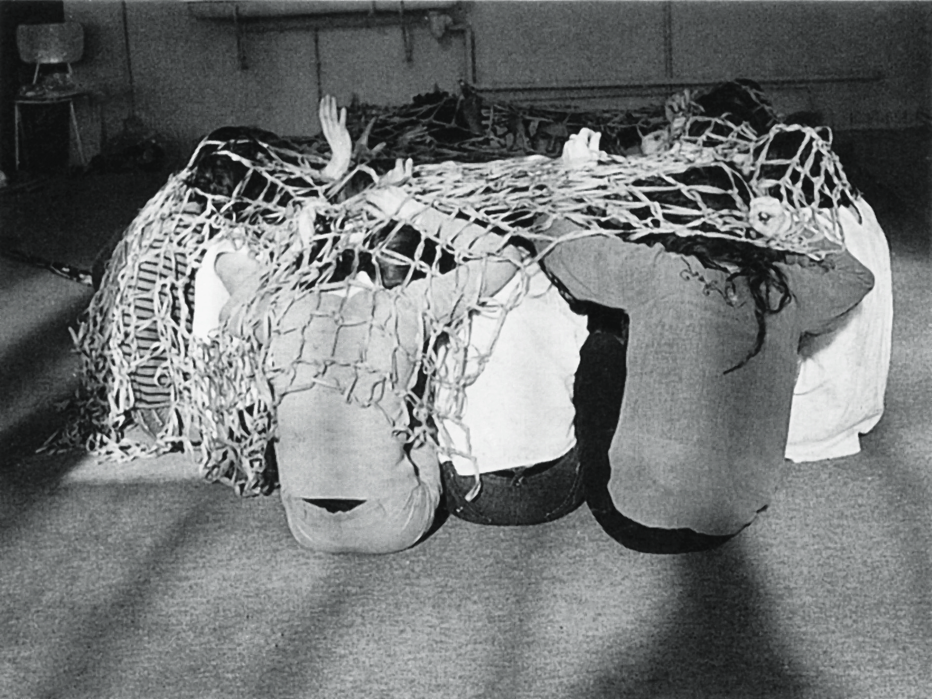 A sepia toned image showing several people sitting on the ground, their knees to their chests. Covering them is net, their hands peak through the holes. 