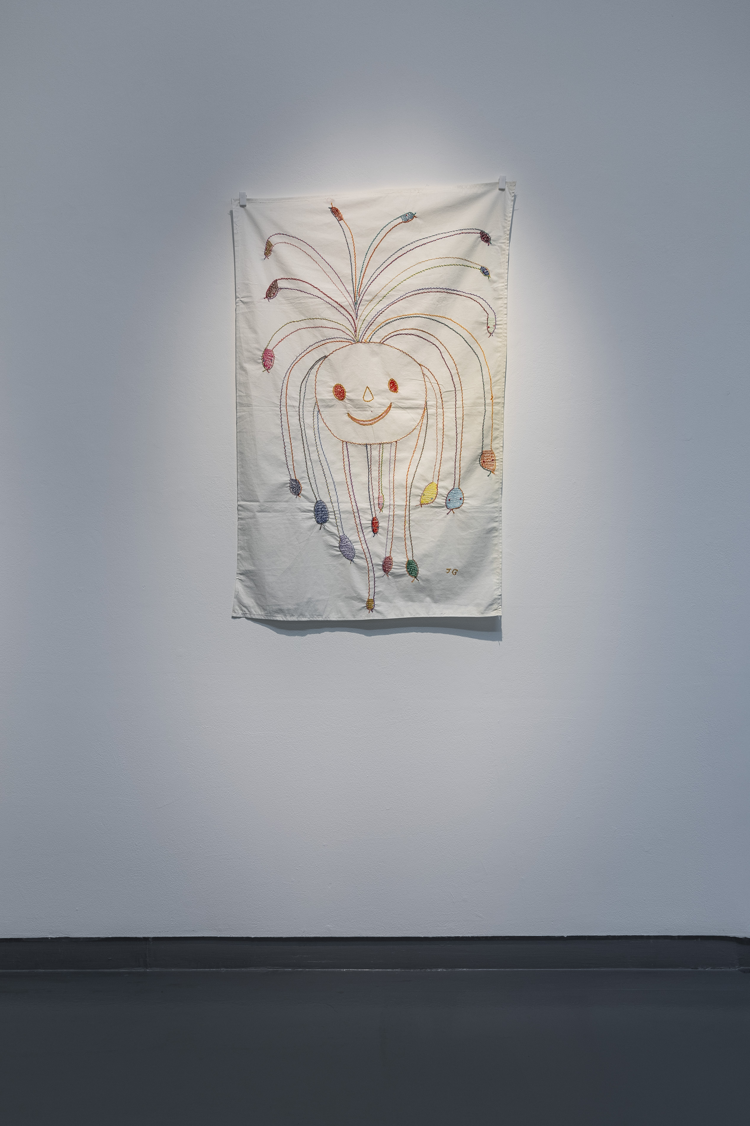 An embroidered fabric work by James Gladwell showing a smiling, round-headed figure with long tendril-like hair, each strand ending in a small coloured bead or creature.
